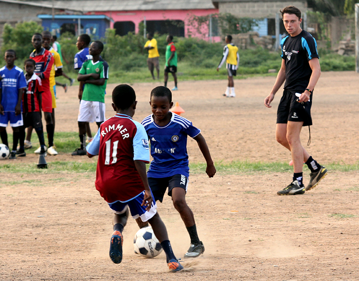 Children playing football on dusty pitch in Ghana with UK coach.